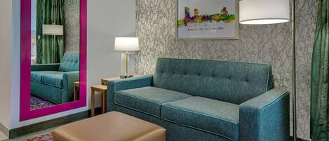 A hotel room with a teal sofa, a brown ottoman, a floor lamp, a mirror, and a framed cityscape picture on the wall.