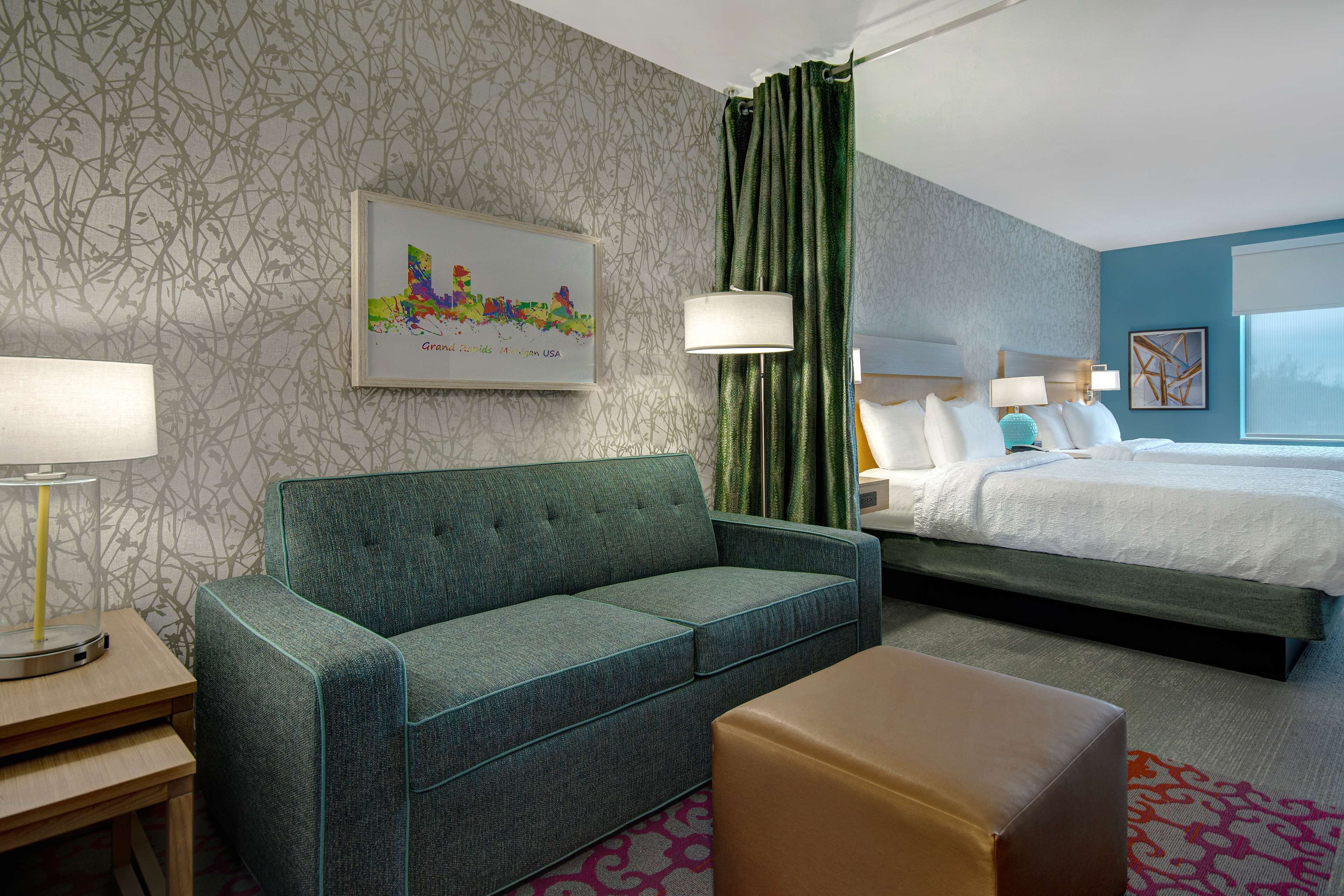 A hotel room with a green sofa, a bed with white bedding, a small table with a lamp, and a colorful cityscape artwork on the wall.