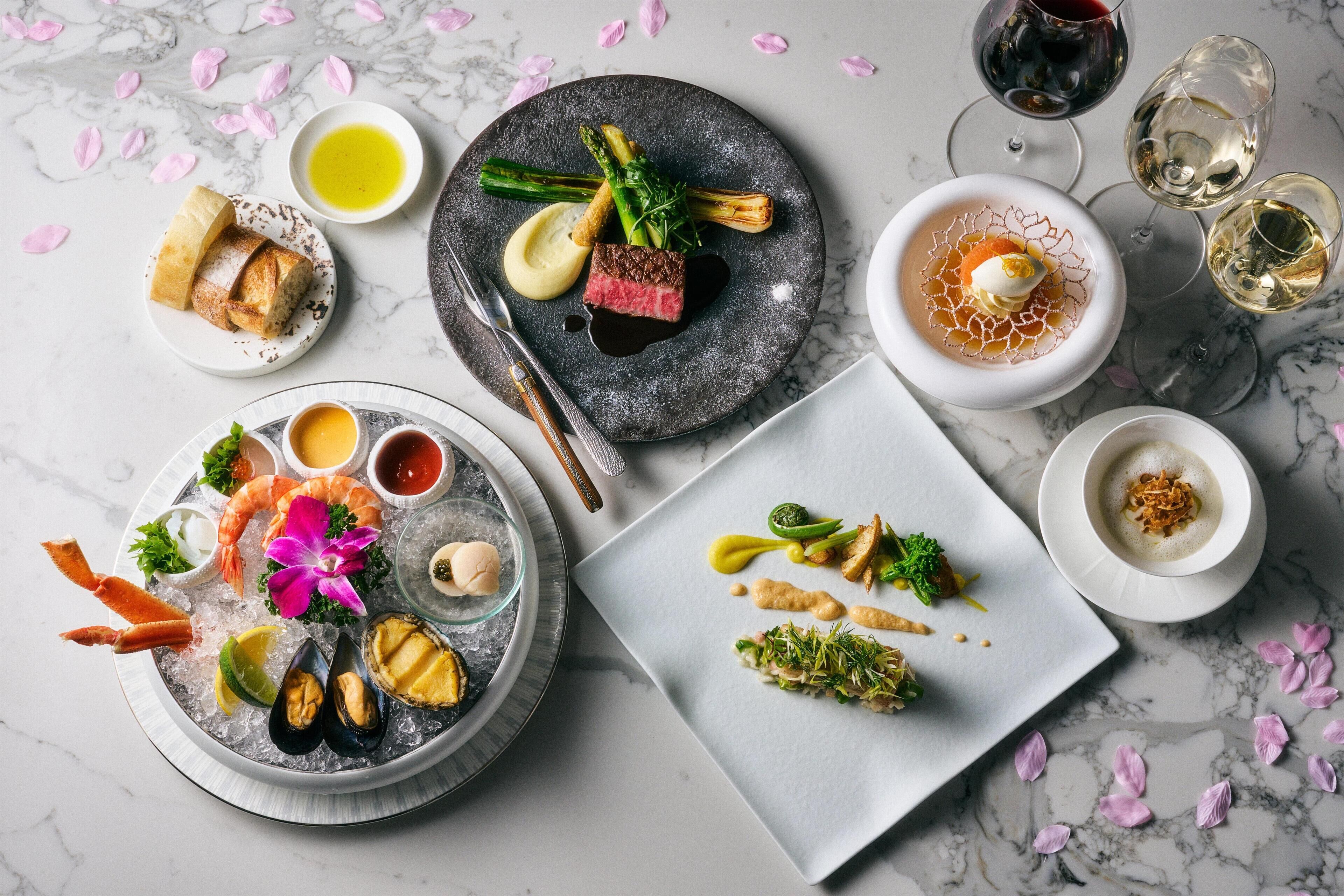 A variety of dishes on plates, including seafood, vegetables, and a steak, with wine glasses and a dessert on the side.
