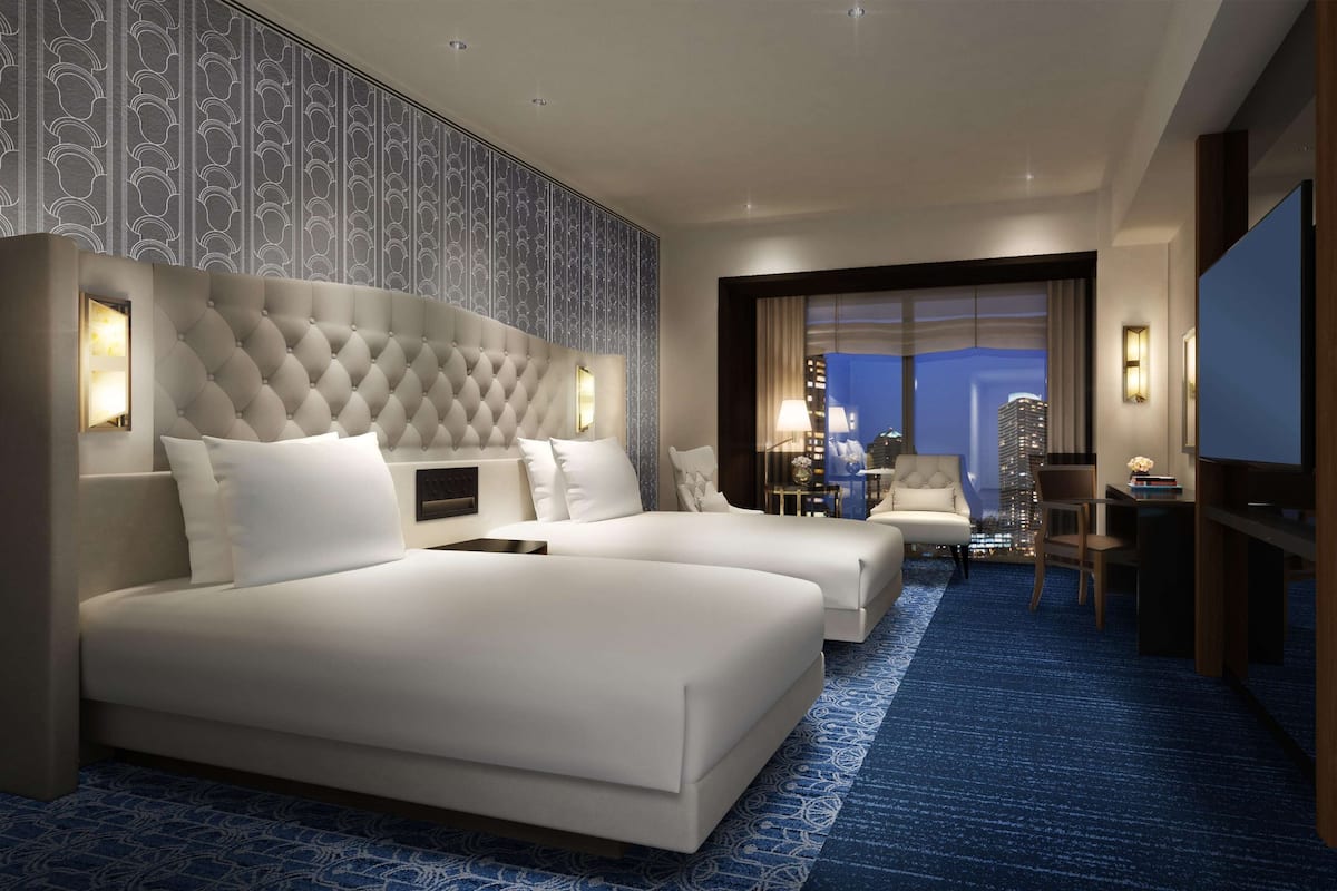Quarto de hotel com uma cama grande, duas camas menores, uma mesa, uma televisão e vista para a cidade.