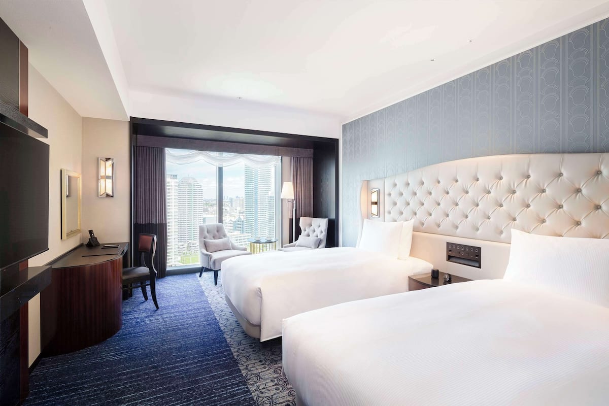 Quarto de hotel com duas camas, uma mesa, uma cadeira e uma janela grande com vista para a cidade.