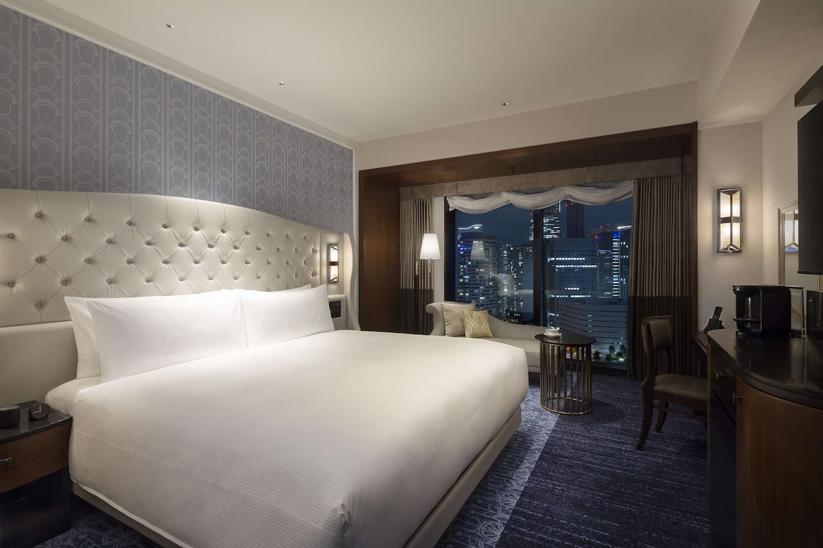 Quarto de hotel com uma cama grande, uma escrivaninha, uma cadeira e vista para o horizonte noturno.