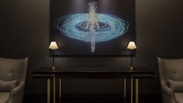 A modern room with a large abstract painting on the wall, a dark console table with two lamps, and two upholstered chairs.