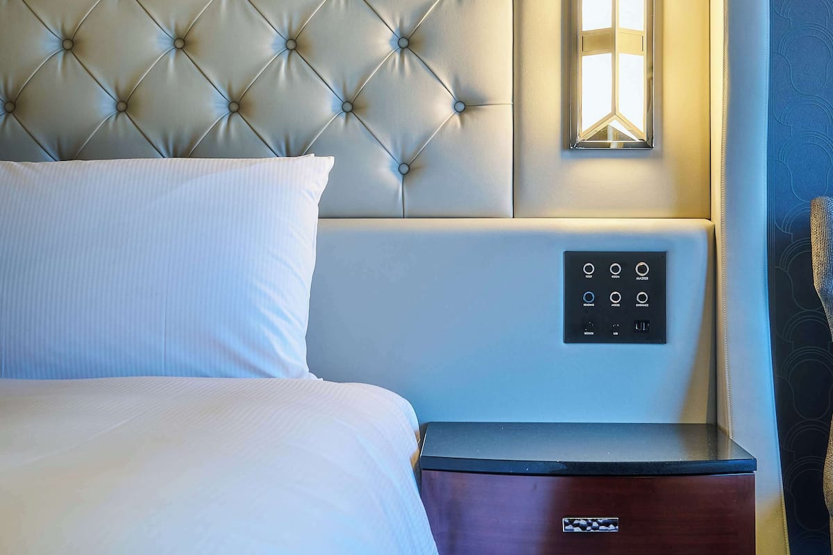 Uma cama bem arrumada com cabeceira estofada, mesa de cabeceira com painel de controle e papel de parede estampado.