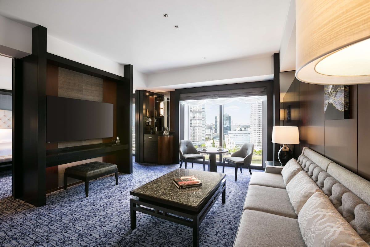 Quarto de hotel moderno com televisão de tela plana grande, um sofá, uma mesa de centro e uma área de jantar com cadeiras.