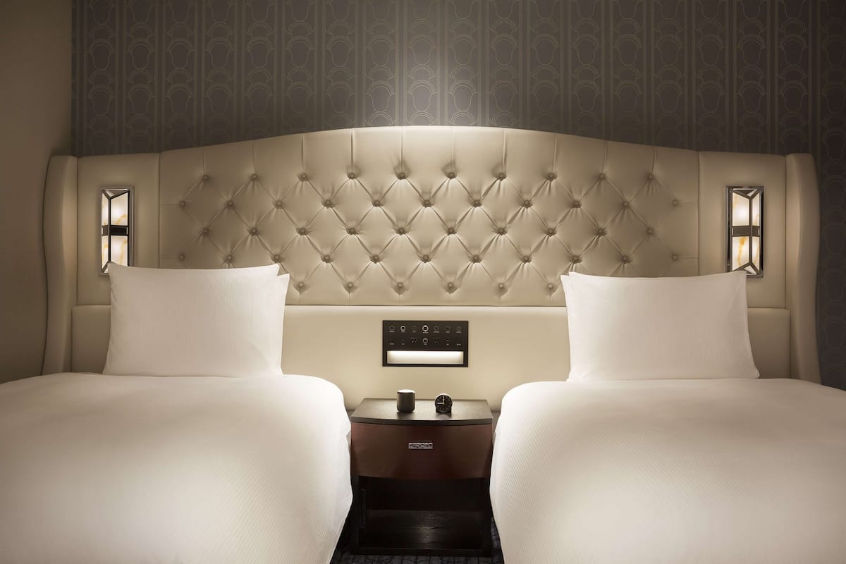 Quarto de hotel com cabeceira estofada, duas camas, mesa de cabeceira com relógio e painel de controle.