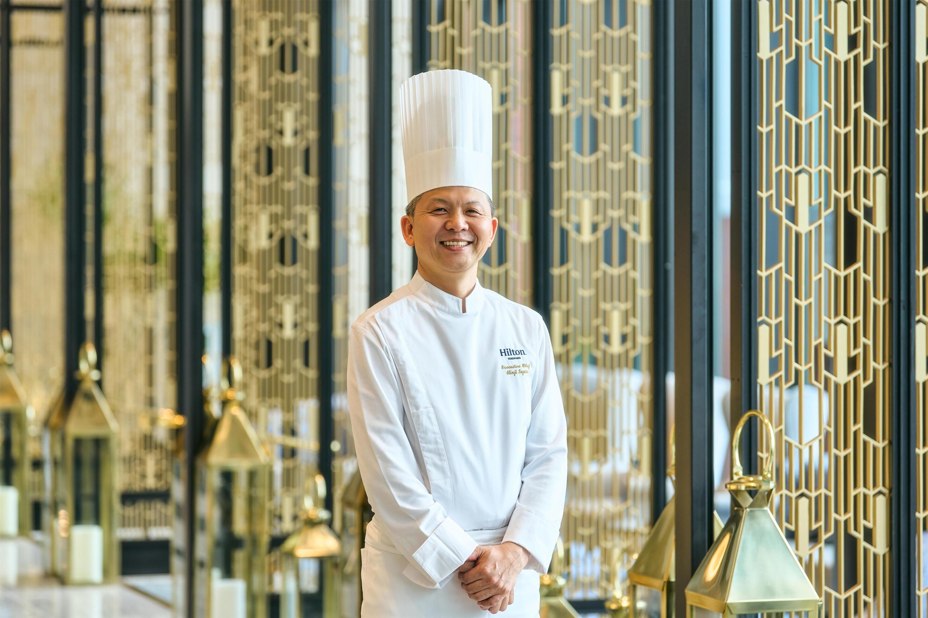 A person in a chef's uniform standing in front of a decorative interior with patterned metalwork.