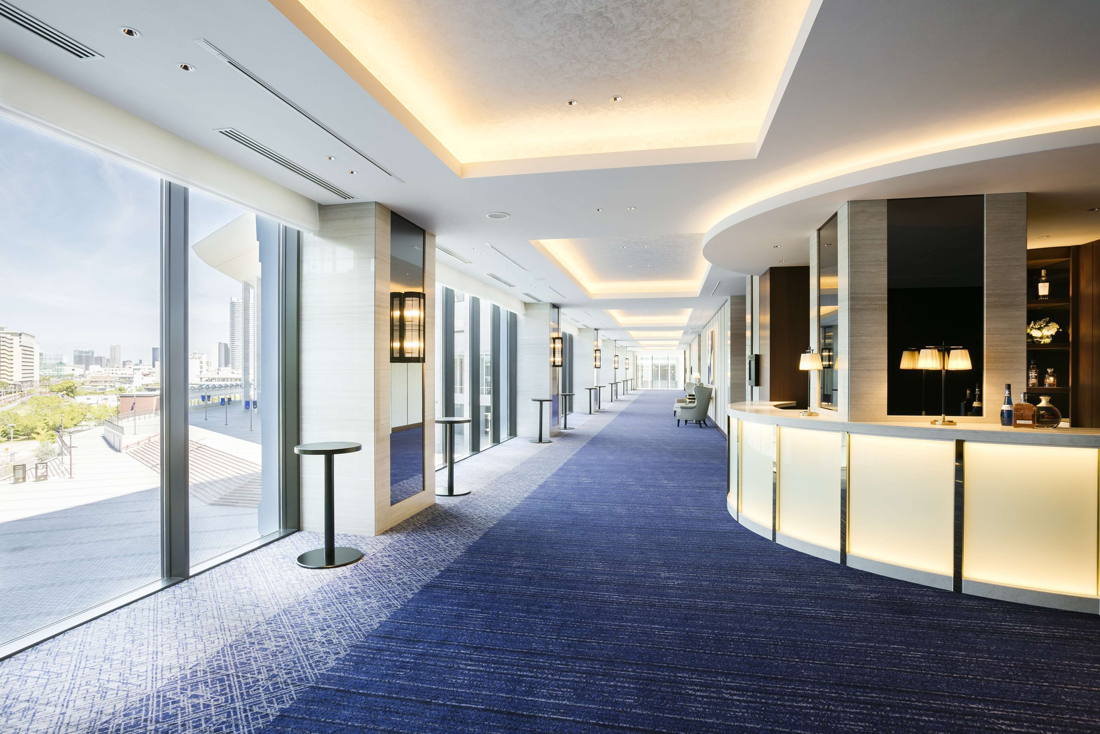 A modern hotel lobby with a blue carpet, glass walls, and a reception desk with a counter and lamps.