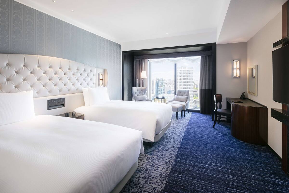 Quarto de hotel com duas camas, uma janela ampla com vista para a cidade, uma escrivaninha e uma cadeira.