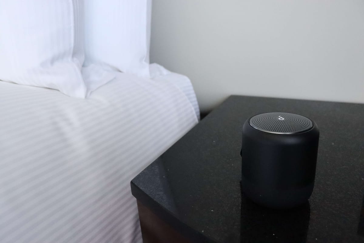 Um alto-falante preto sobre uma pequena mesa preta ao lado de uma cama com roupa de cama branca.