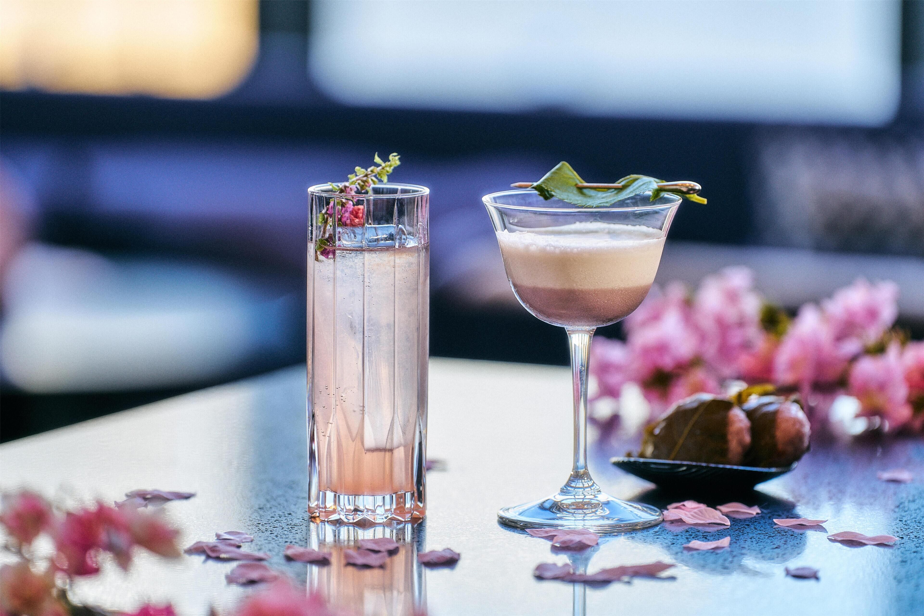 Two cocktails on a table with rose petals and a bowl of fruit.