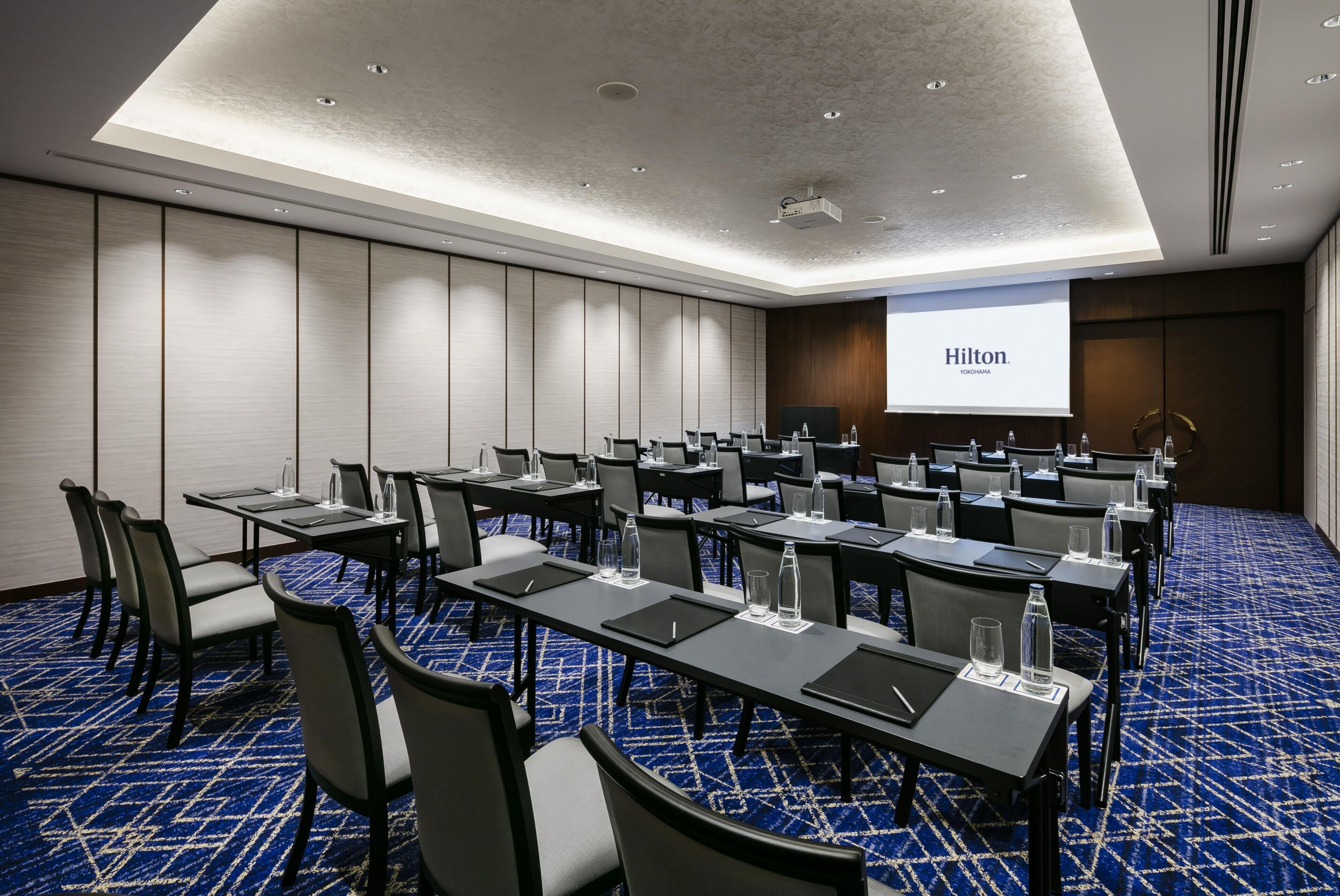 A conference room with multiple tables and chairs, a large screen displaying 'Hilton Yokohama', and a blue patterned carpet.