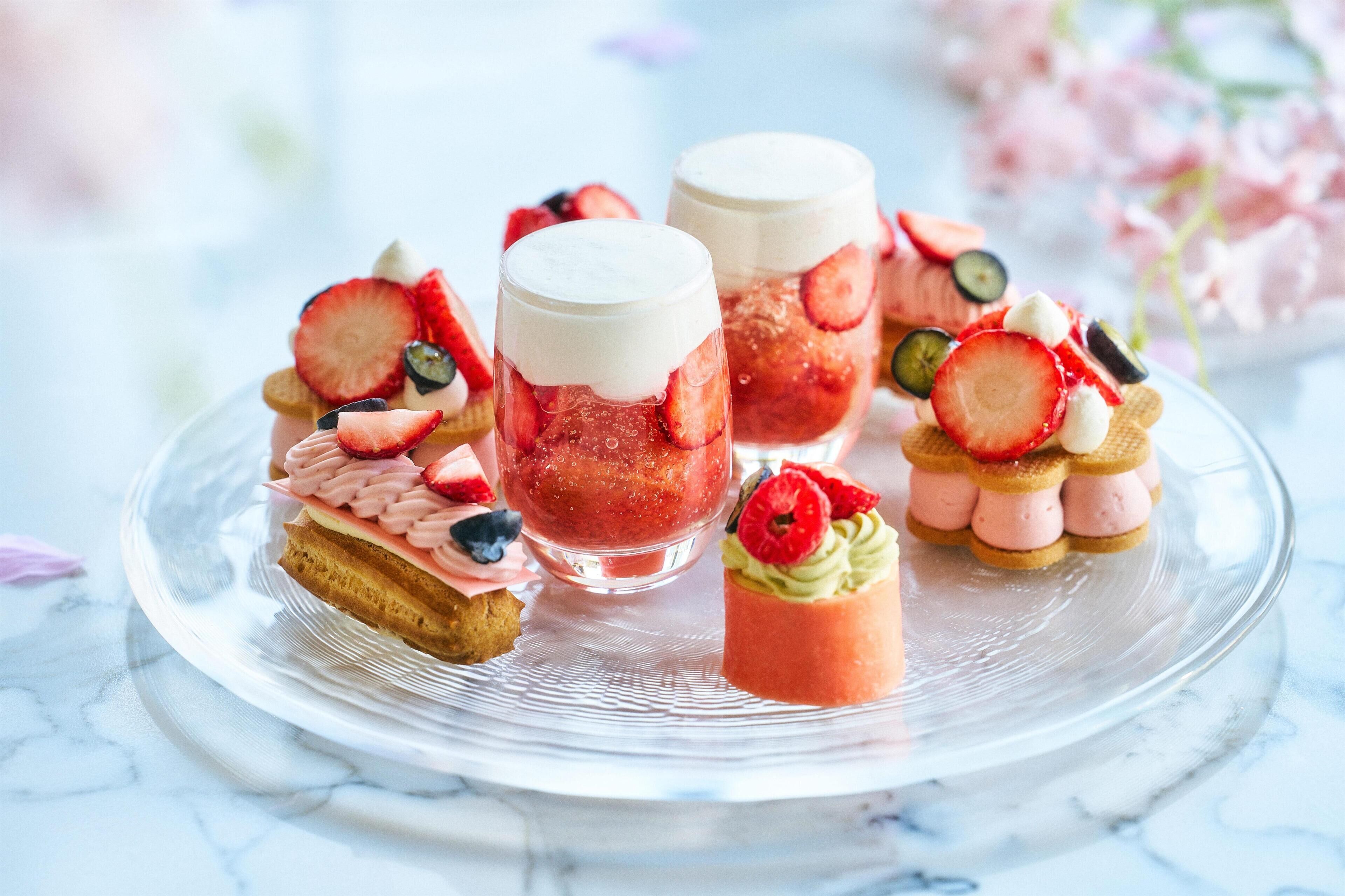A variety of desserts on a clear glass plate, including a layered cake, a parfait, and small pastries with fruit toppings.