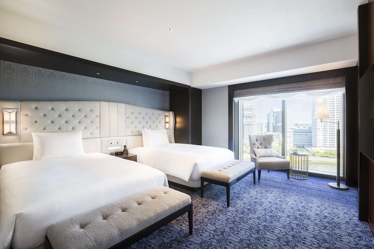Quarto de hotel moderno com duas camas, uma janela grande com vista para a cidade e uma varanda com cadeira e mesa.