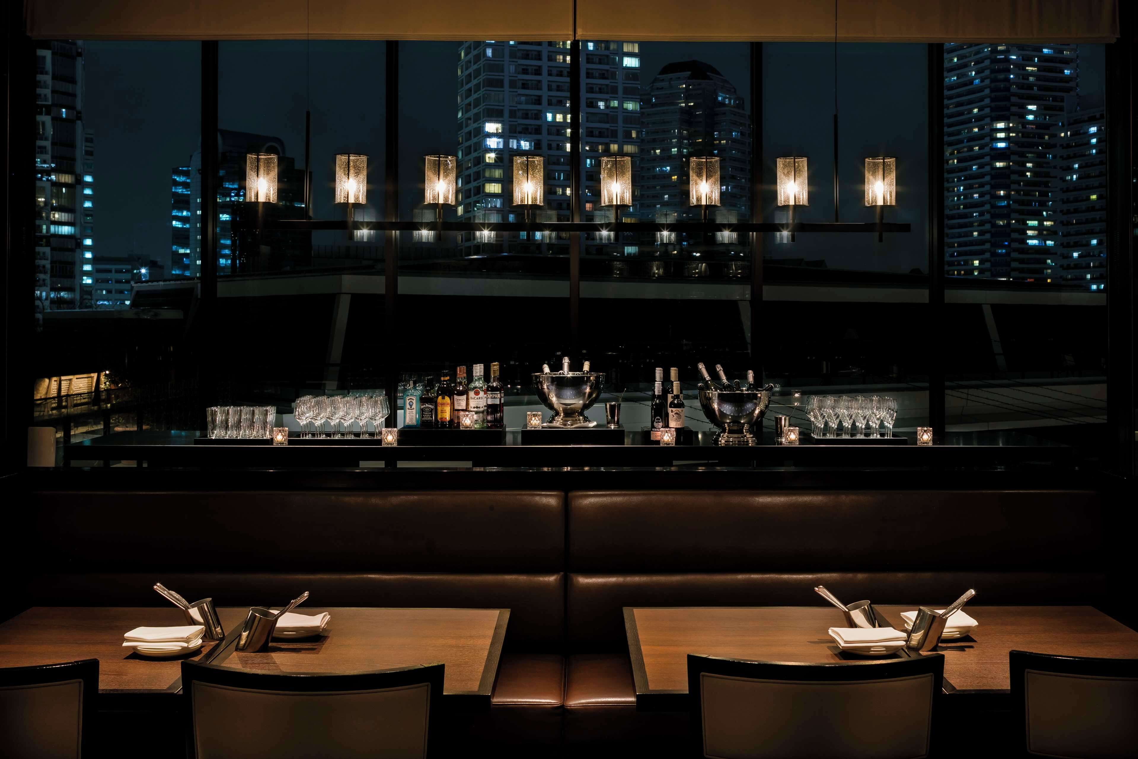 A restaurant with a city view, tables set with cutlery and glasses, and a bar area with bottles and glasses.