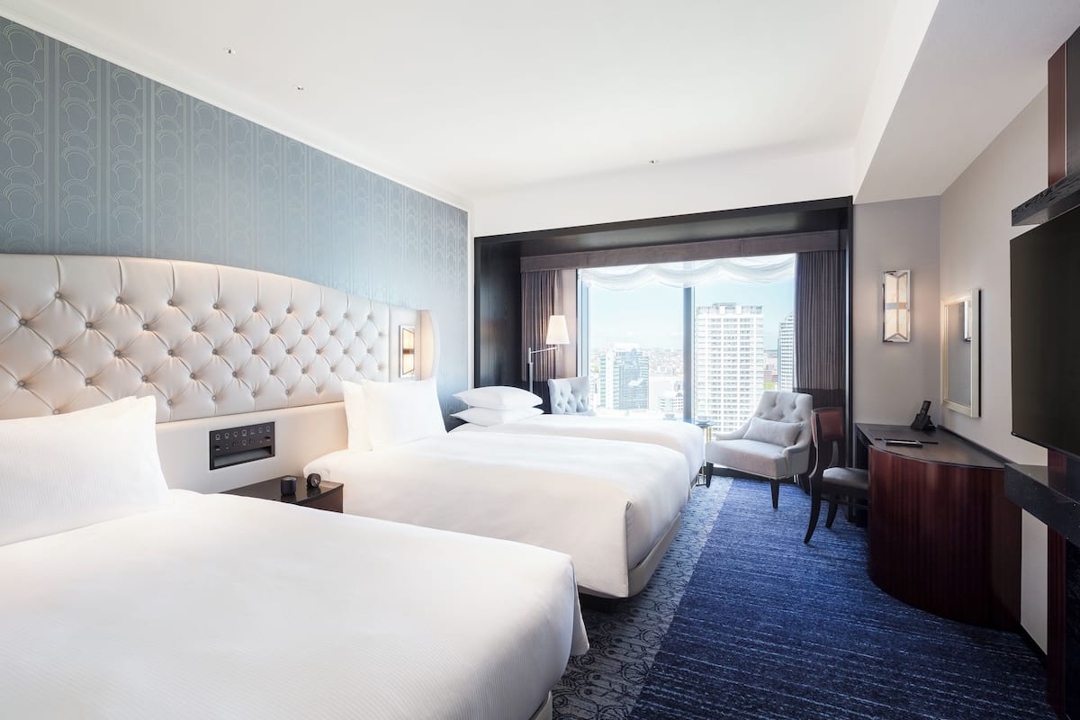 Quarto de hotel com uma grande cabeceira estofada, duas camas, uma escrivaninha com cadeira e uma janela ampla com vista para a cidade.