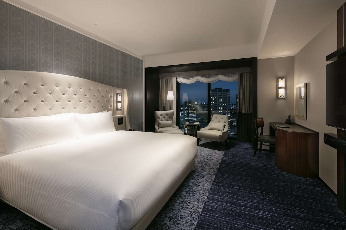 Quarto de hotel com uma cama grande, uma escrivaninha, uma cadeira e vista para a cidade à noite.