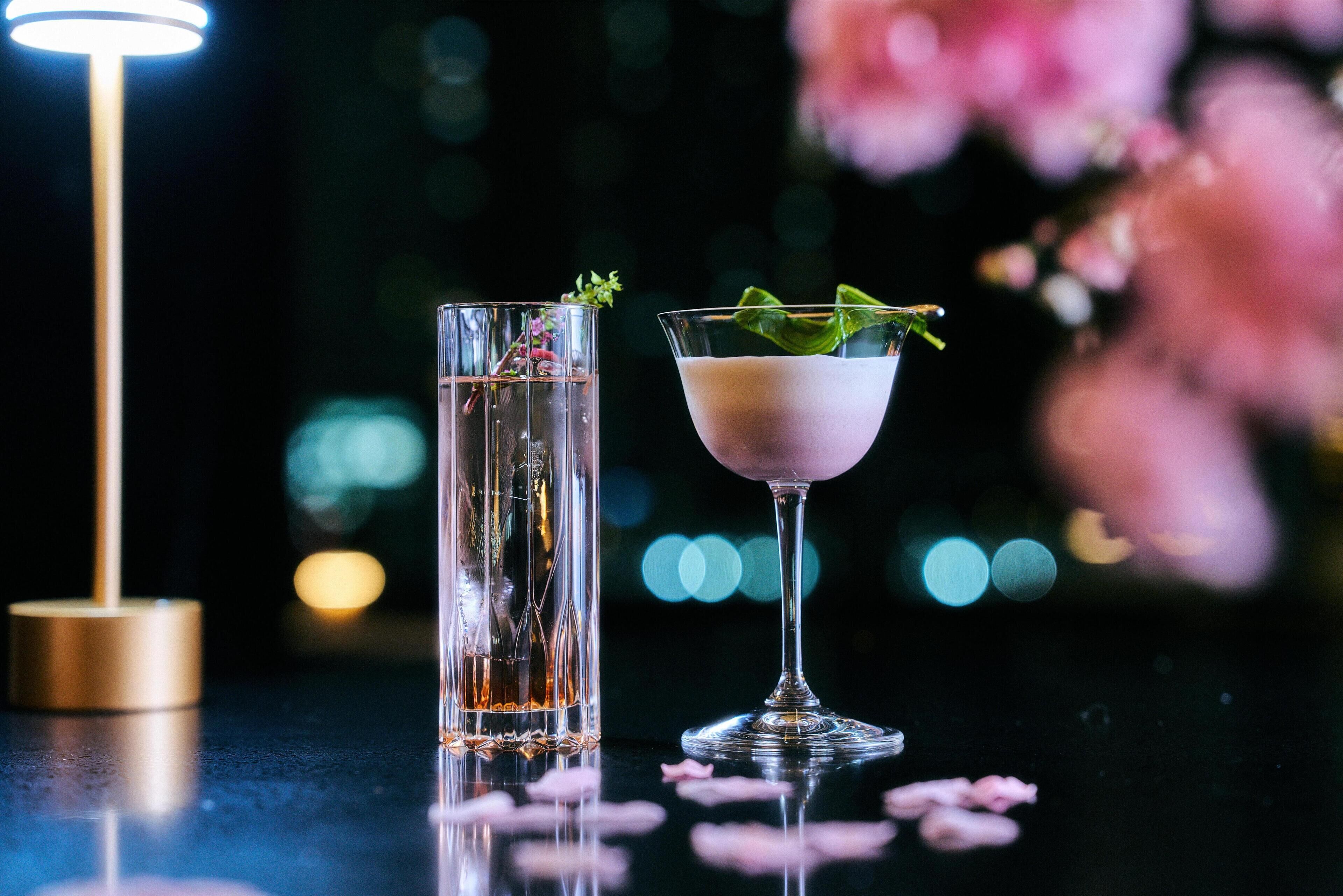 Deux boissons posées sur une surface réfléchissante, avec un arrière-plan flou composé de lumières et de fleurs roses.