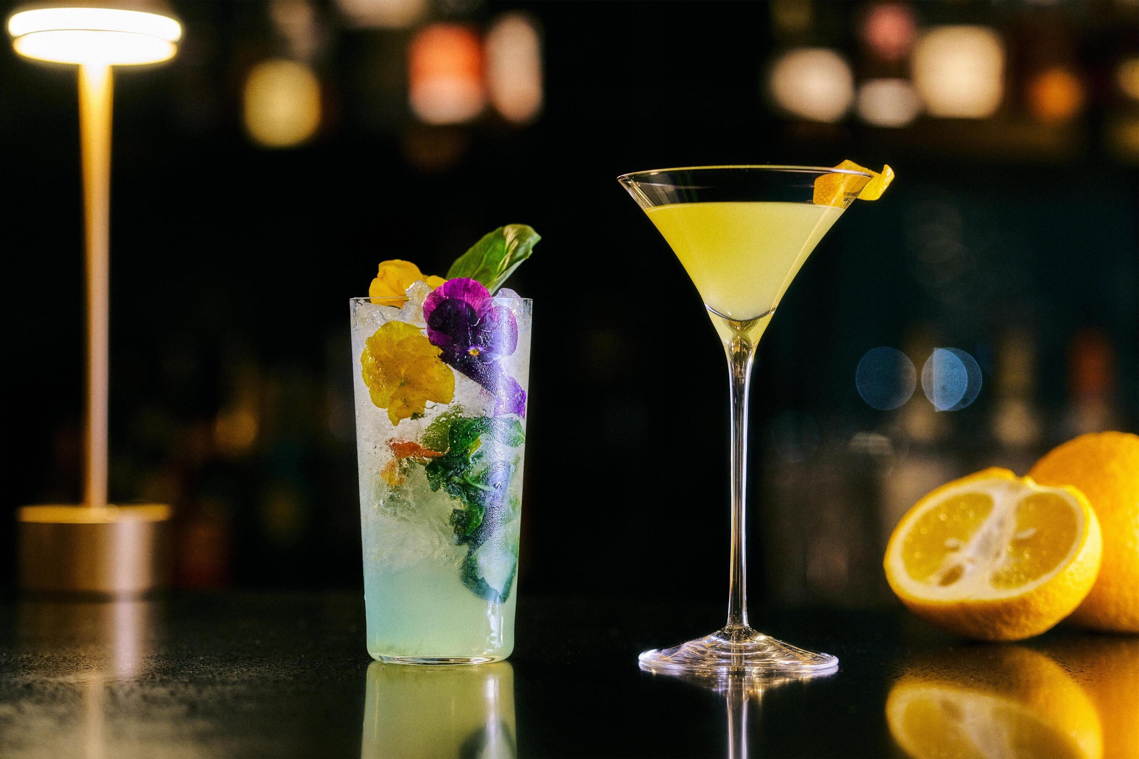 Two drinks on a bar counter, one in a tall glass and one in a martini glass, with a lit lamp in the background.