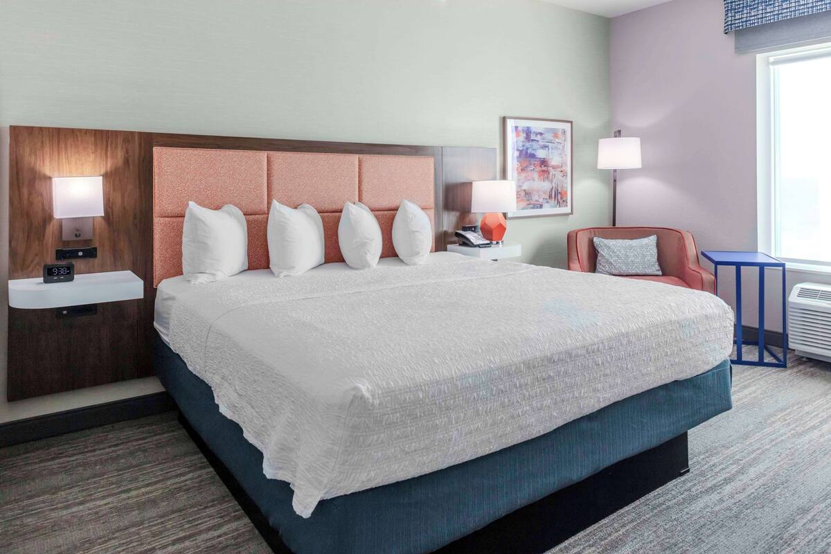 A hotel room with a large bed, a nightstand with a lamp, a chair, and a window with a view.