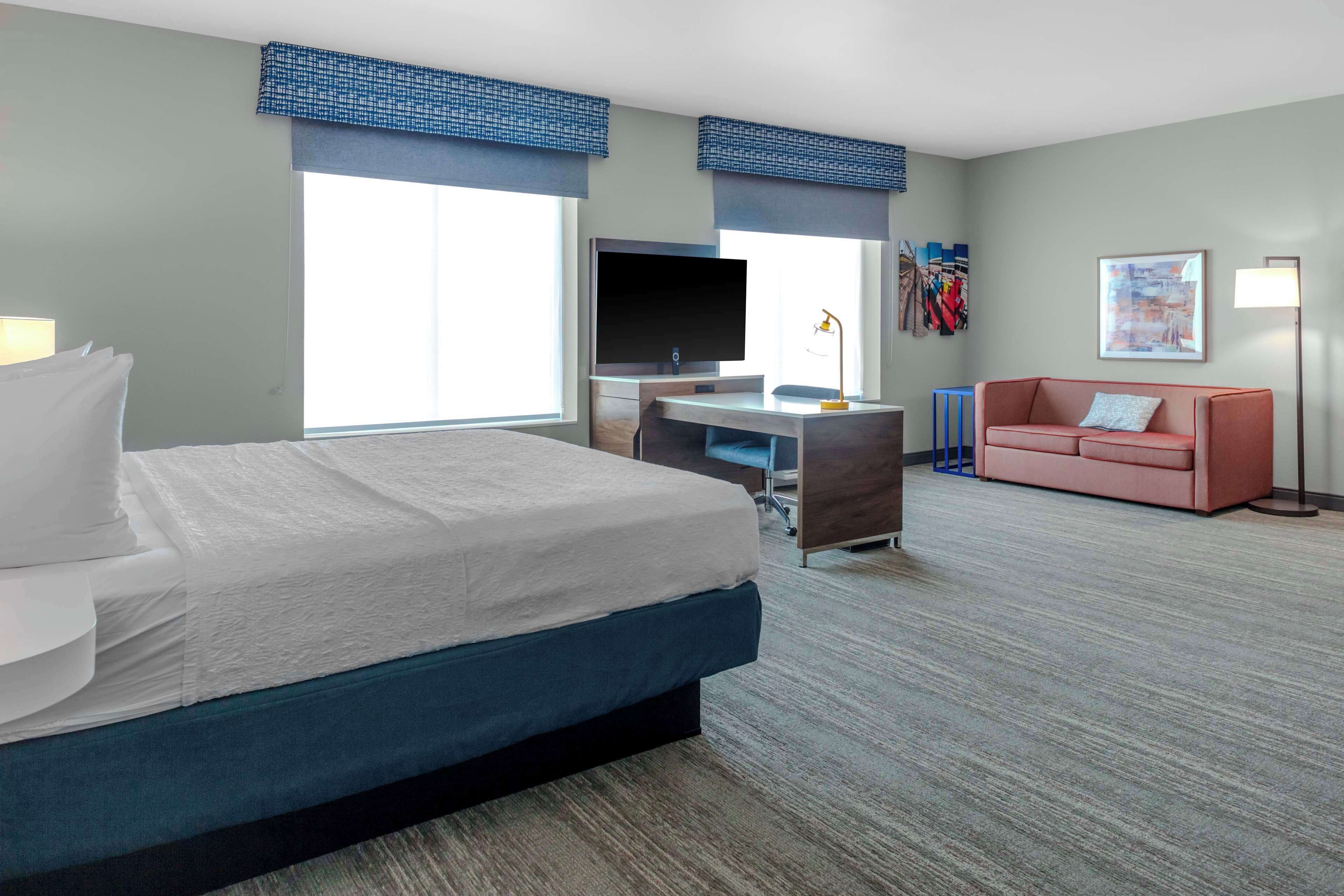 A hotel room with a bed, a desk with a TV, a pink sofa, and large windows.