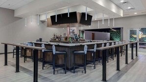 A modern bar area with a long counter, multiple stools, and a variety of bottles. - Hampton Inn & Suites Indianapolis West Speedway (Indianapolis)