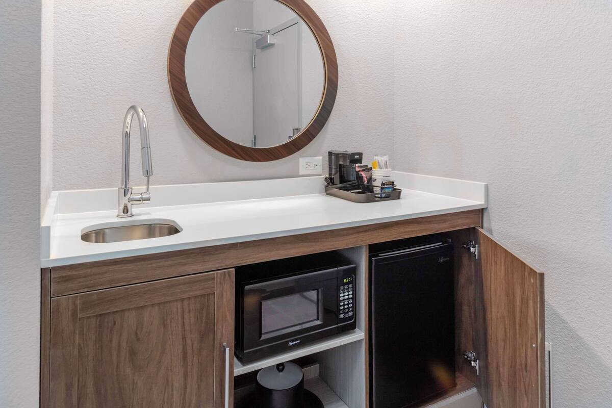A modern bathroom with a white sink, a round mirror, a built-in microwave, and a black refrigerator.