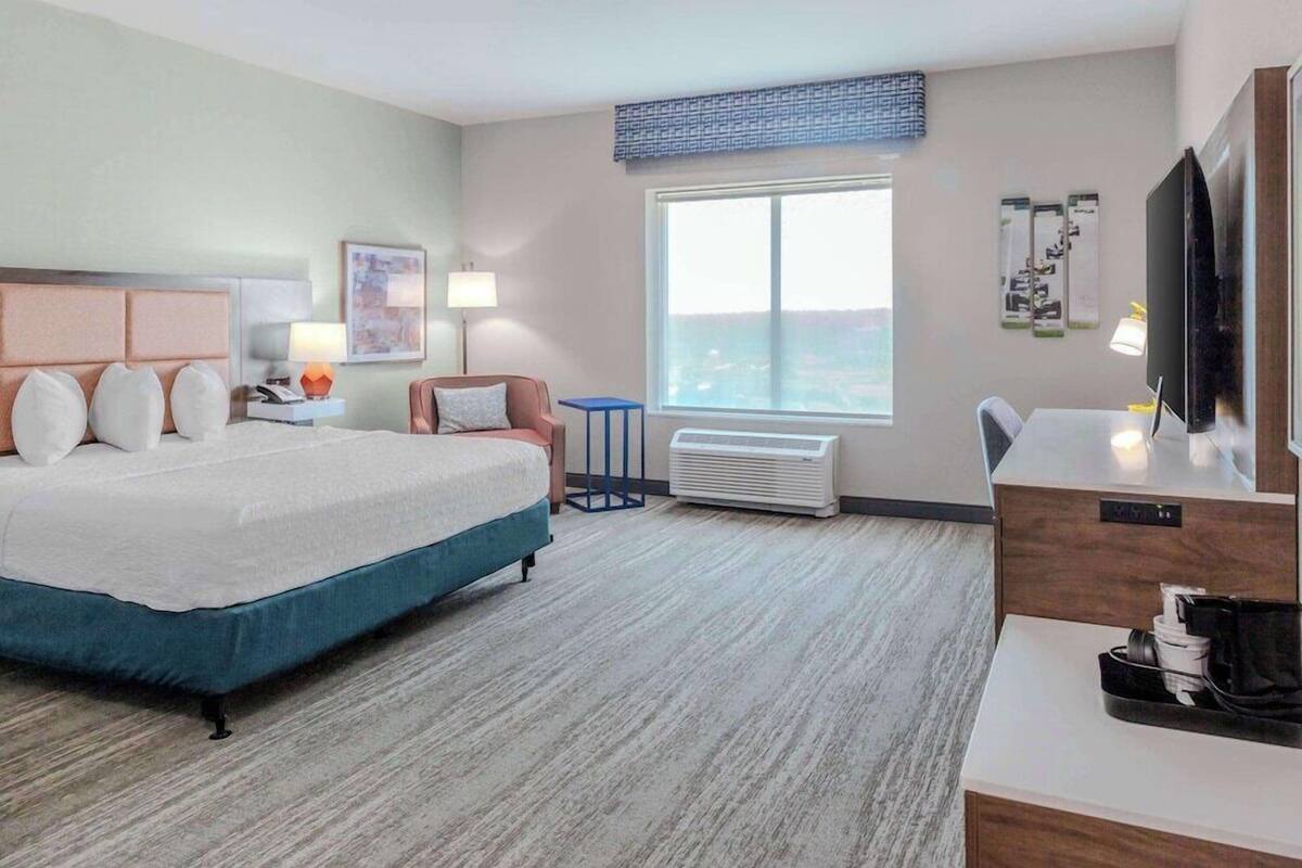 A hotel room with a bed, a desk, a chair, a TV, a window with a view, and an air conditioning unit.