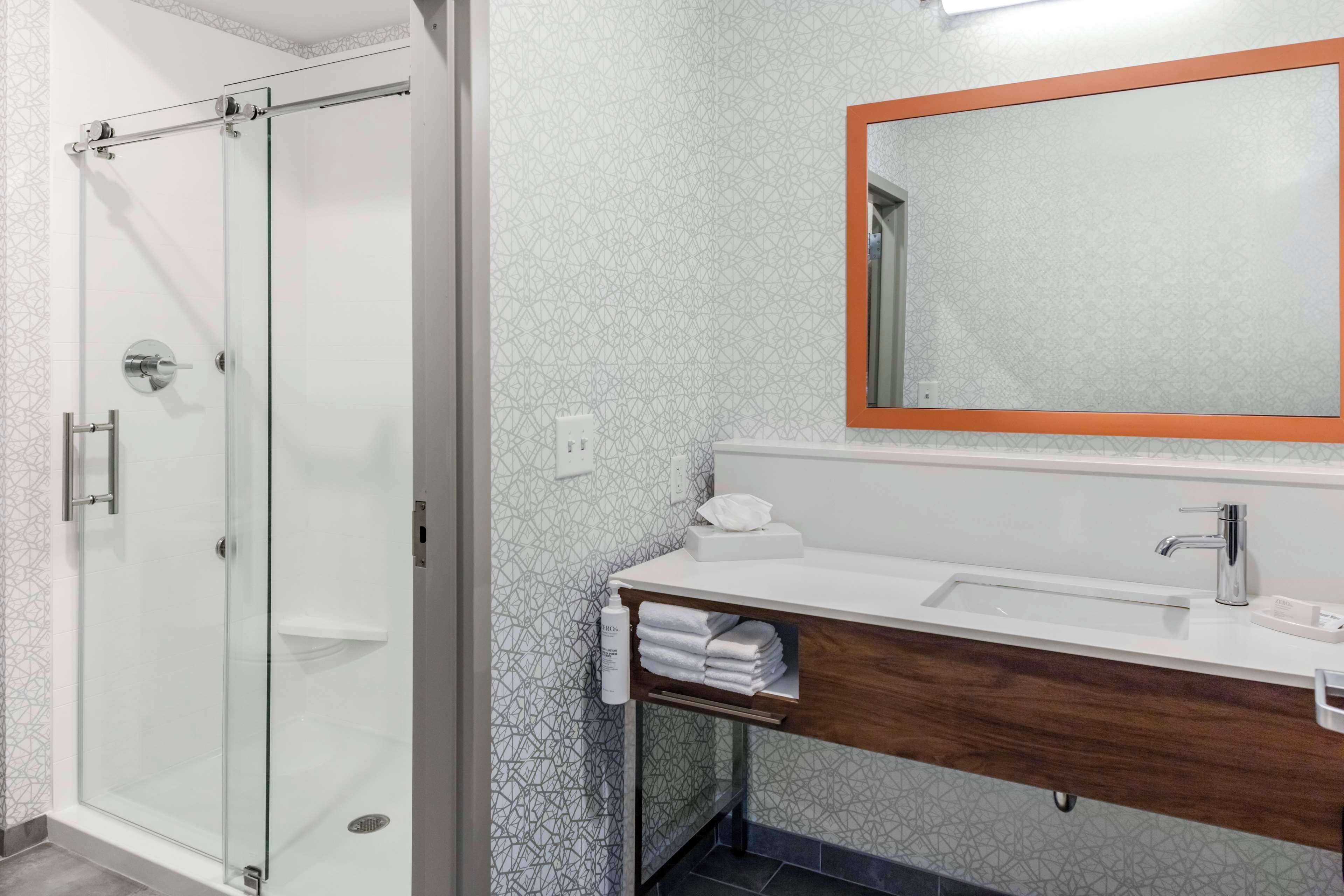 A modern bathroom with a glass shower enclosure, a white sink with a wooden vanity, and a large mirror with an orange frame.