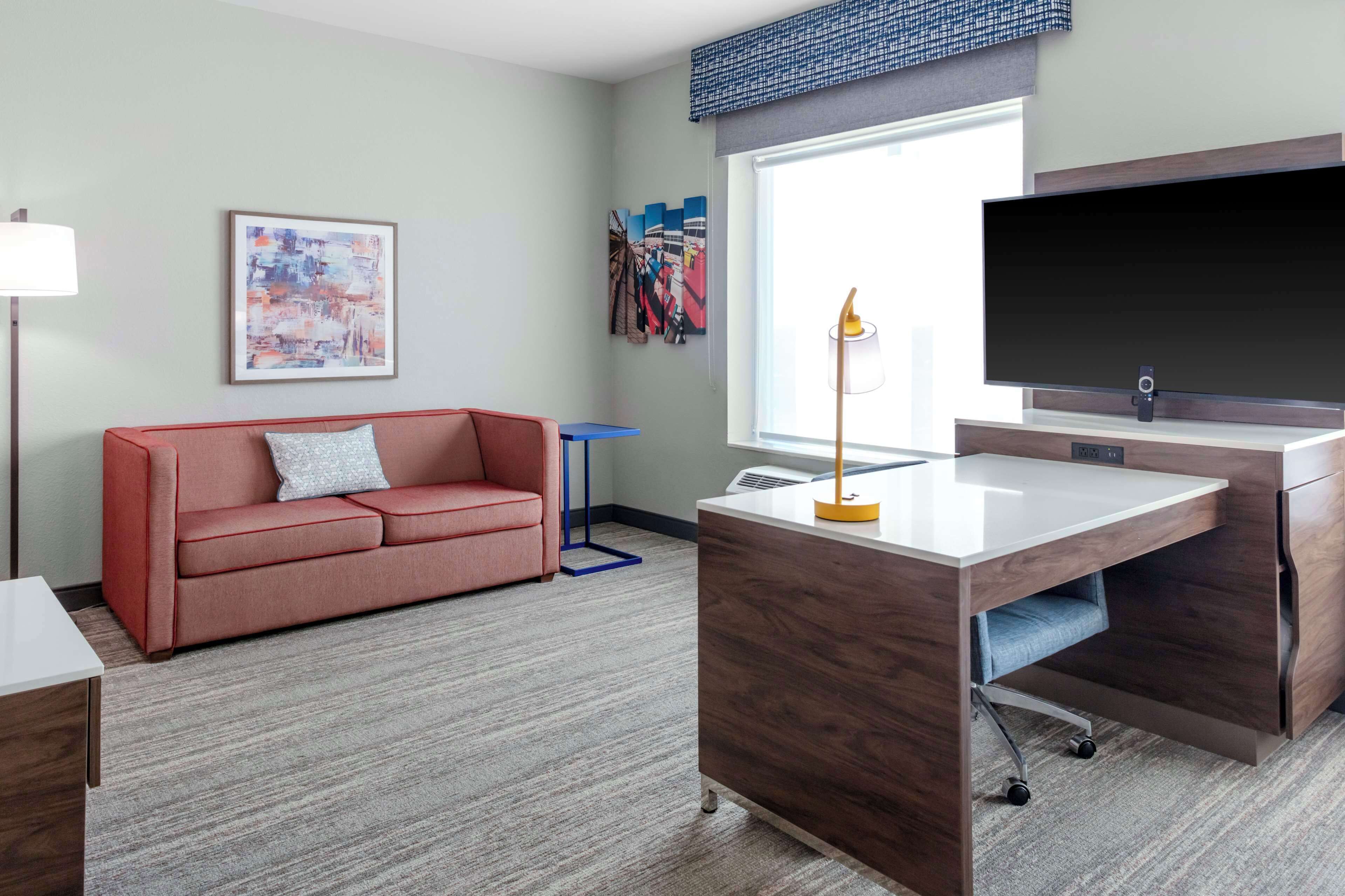 A modern hotel room with a red sofa, a desk with a flat-screen TV, and a large window providing a city view.
