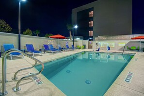 Une piscine entourée de chaises longues bleues et de parasols rouges, le tout bordé par un bâtiment doté de nombreuses fenêtres.