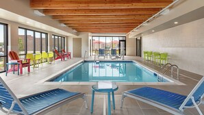 Une piscine avec des chaises longues et des chaises colorées, des poutres en bois au plafond et une vue sur l’extérieur.
