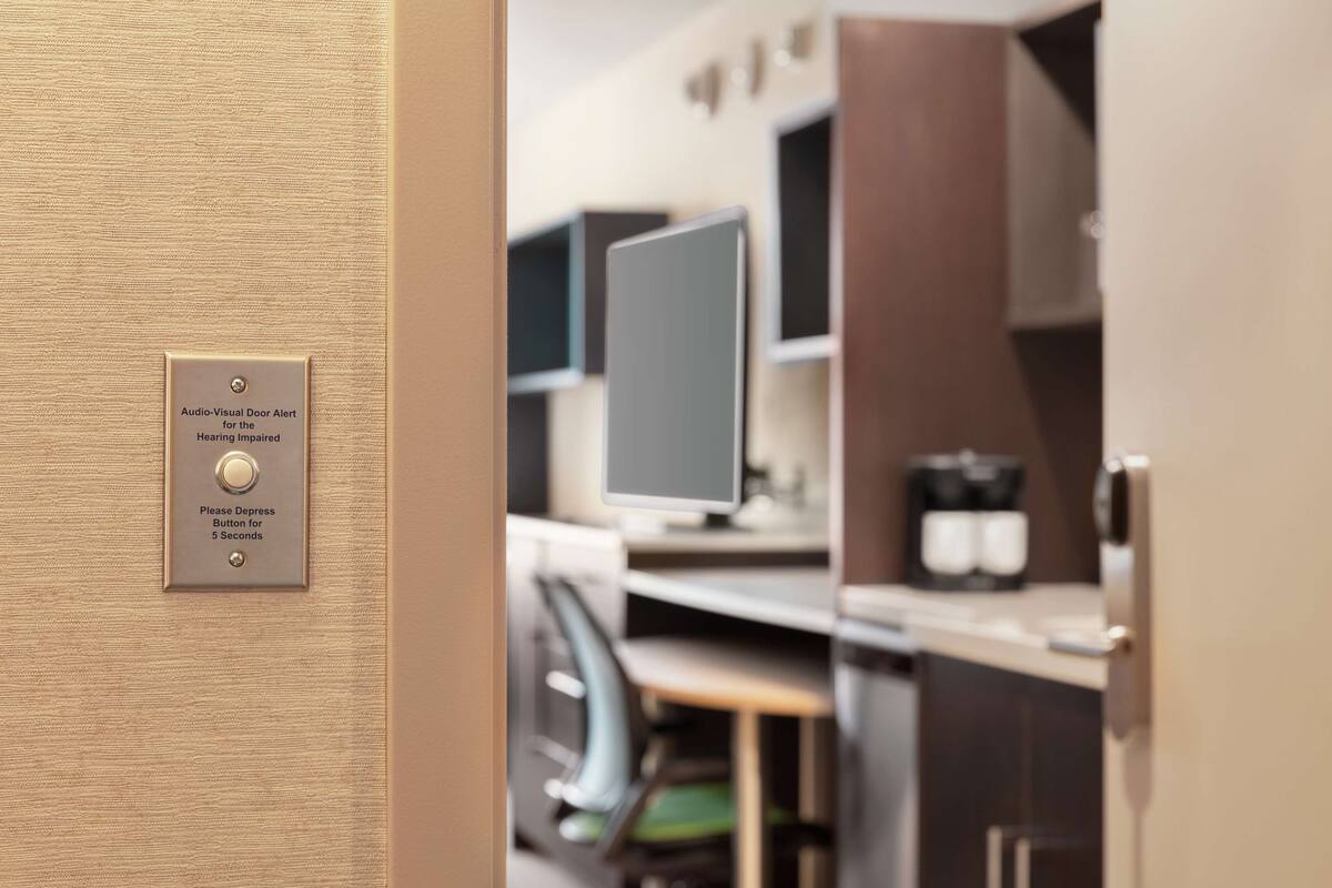 Een deur met een audiovisueel alarmsysteem voor mensen met een gehoorbeperking, een knop die 5 seconden ingedrukt moet worden, en een modern interieur met een flatscreenmonitor en keukenapparatuur.