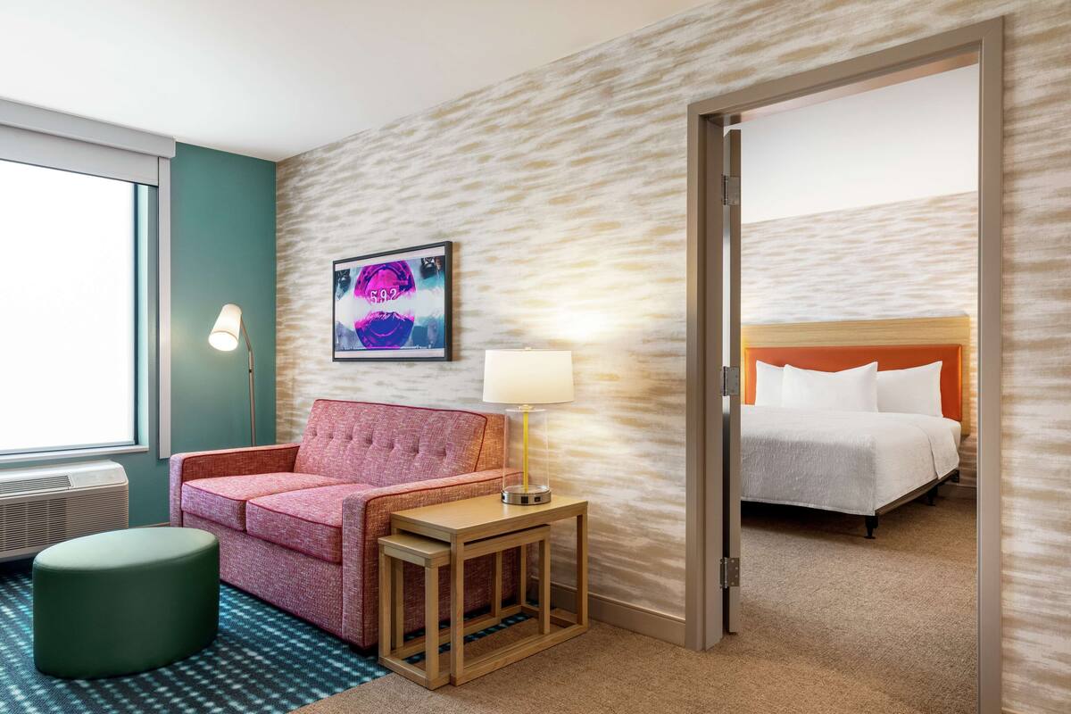 Een hotelkamer met een bed, een bank, een kleine tafel met een lamp, een kunstwerk aan de muur en een groot raam.