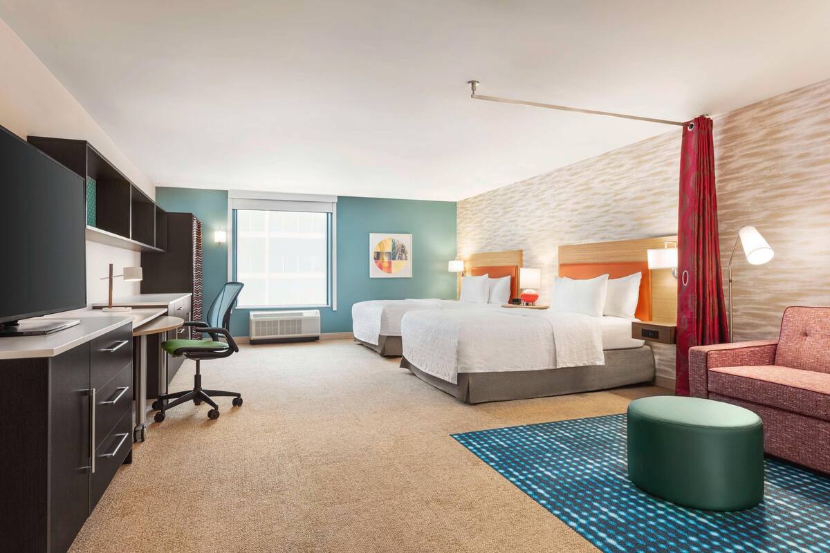 Une chambre d’hôtel avec un bureau, deux lits, une chaise, une lampe et une fenêtre.