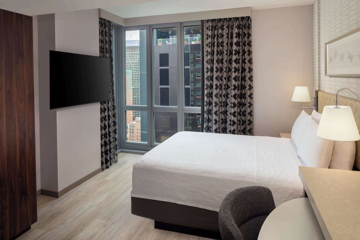 Une chambre d’hôtel avec un grand lit, un téléviseur fixé au mur, un bureau avec une lampe et une vue sur de hauts immeubles par la fenêtre.