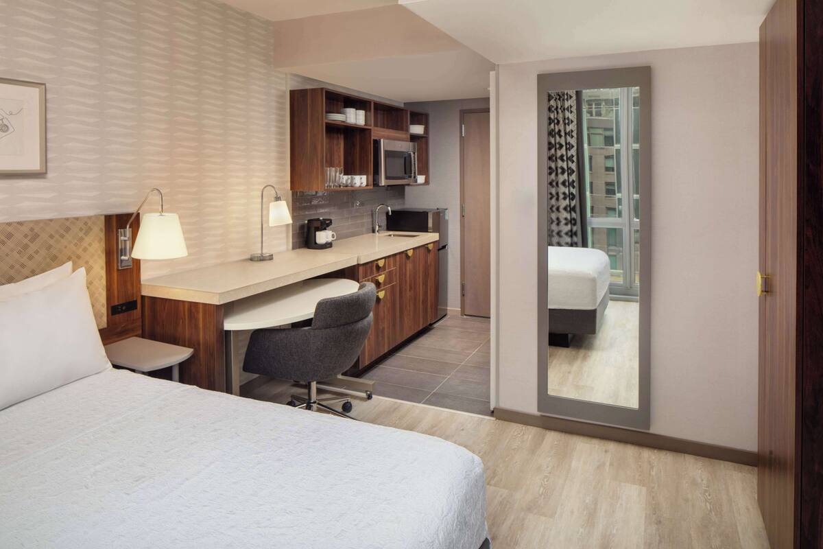 Une chambre d’hôtel comprenant un lit, un bureau avec une chaise, une kitchenette et une porte coulissante donnant sur un balcon.