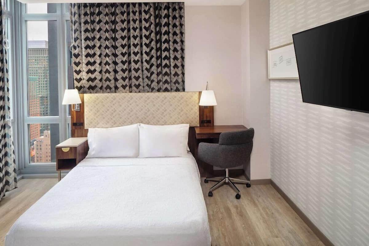 Une chambre d’hôtel avec un grand lit, un bureau avec une chaise, une télévision et une fenêtre avec des rideaux.