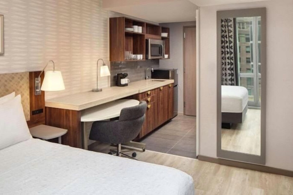 A hotel room with a bed, a desk, a chair, a small table, a microwave, and a large mirror.