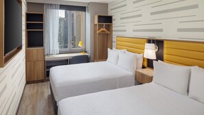 A hotel room with two beds, a desk, a chair, a wardrobe, and a window with a view of buildings.