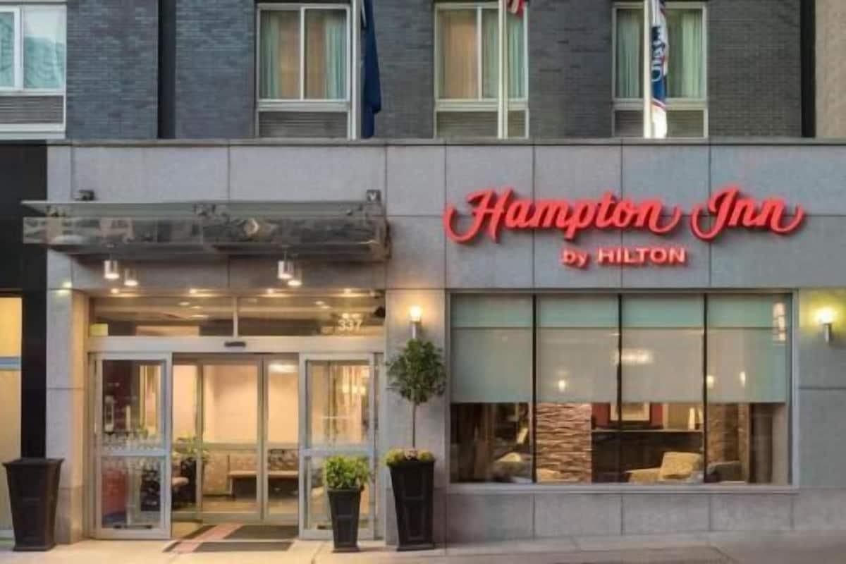 A hotel entrance with a glass facade, a sign reading 'Hampton Inn by Hilton,' and a small outdoor seating area with plants.