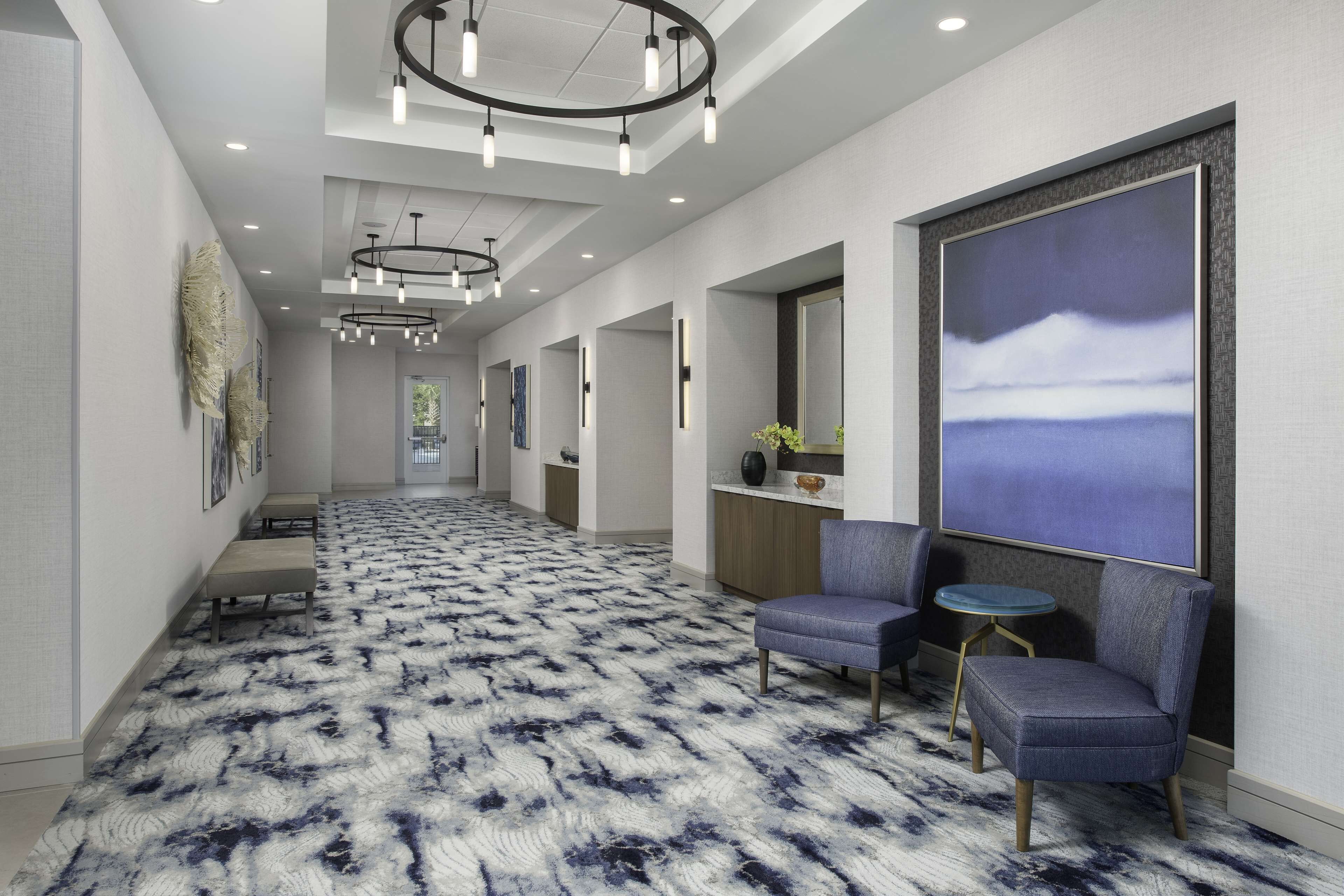 A modern hotel hallway with a patterned carpet, seating area with chairs and a small table, and a large artwork on the wall.