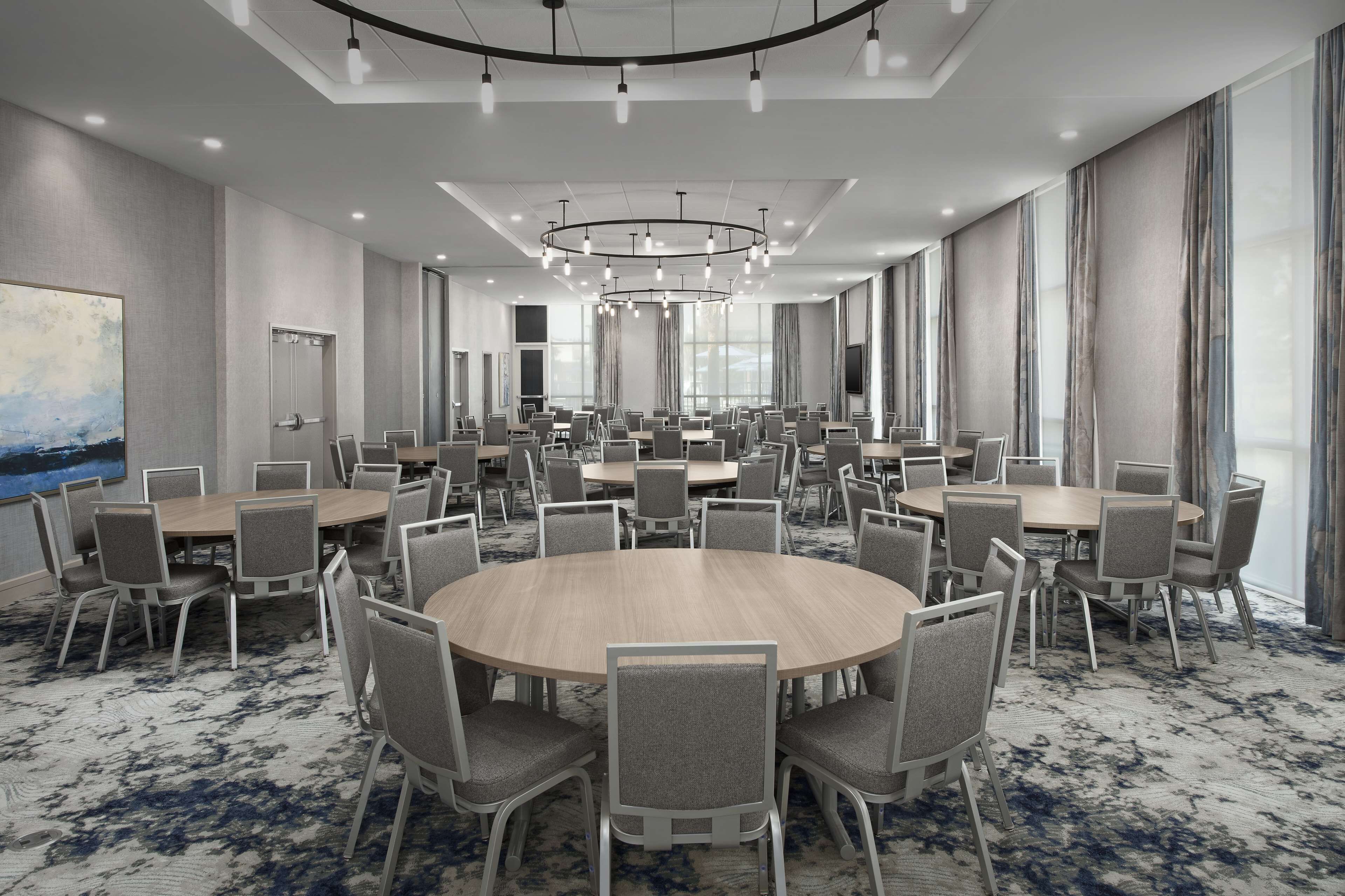 A large conference room with multiple round tables and chairs, a modern chandelier, and large windows with curtains.