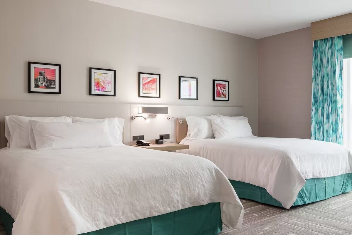 Quarto de hotel com duas camas, uma mesa de cabeceira, uma luminária e uma janela grande com cortinas.