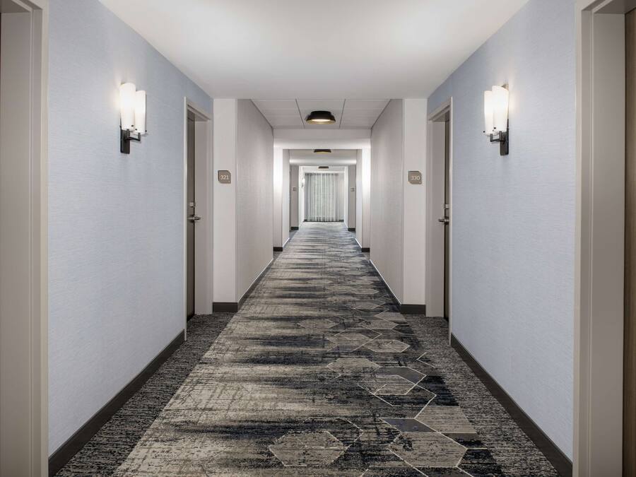A hotel hallway with a carpeted floor, light-colored walls, and wall-mounted light fixtures.