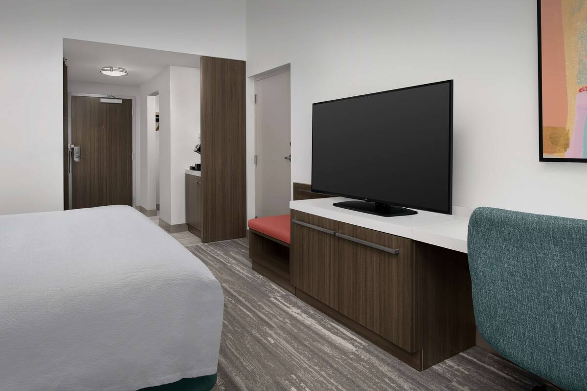 Ein Hotelzimmer mit Bett, Schreibtisch mit Flachbildfernseher, Stuhl und einer Tür zu einem anderen Raum.