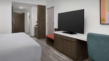 A hotel room with a bed, a desk with a flat-screen TV, a chair, and a door leading to another room.