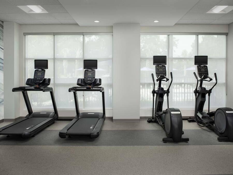 A fitness room with treadmills, elliptical machines, and stationary bikes, large windows with blinds, and a grey carpeted floor.