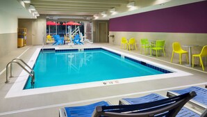 Una piscina interior con marcas claras en los carriles, rodeada de sillas amarillas y verdes, y una pared morada.