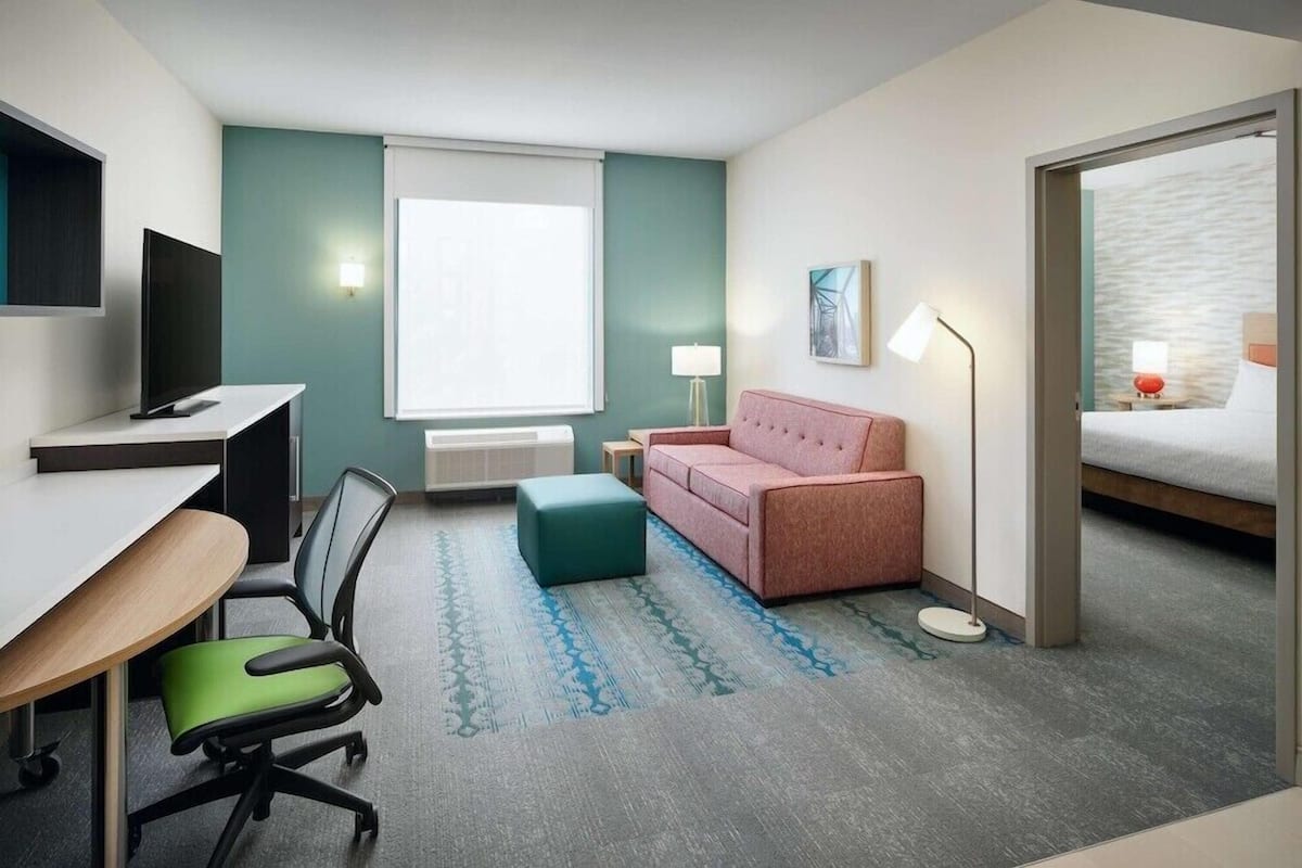 A hotel room with a desk, chair, pink sofa, ottoman, lamp, and a bed with a view.